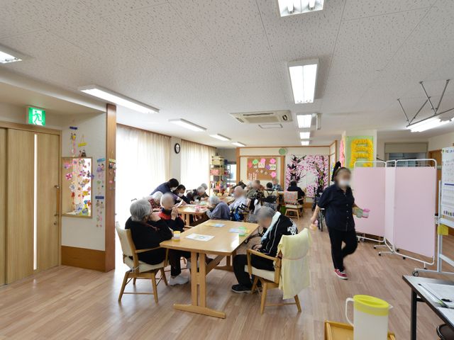 明るい共有スペース リビングルーム2 広々とした部屋で利用者が様々なアクティビティを楽しんでいます。手作業にいそしむ姿や会話を楽しむ様子が見て取れます。