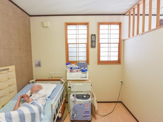 居室の様子。左側には電動ベッドがあり、利用者様が休んでいる。介護、看護用の機材や収納ボックスが置かれている。
