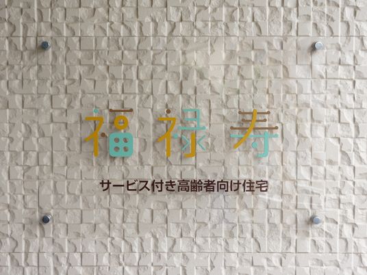 施設の壁には、施設名が表示されている。透明のアクリル板に水色、黄色、茶色の優しい色合いで、施設のロゴが印字されている。