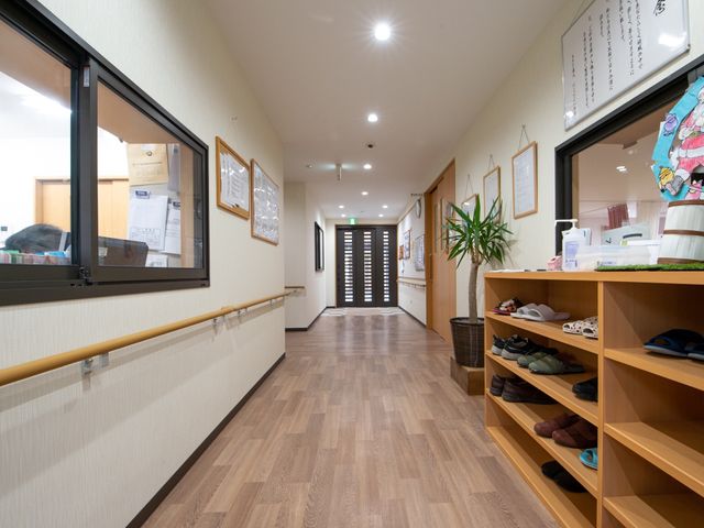 室内の清潔な廊下 玄関 手すりが整備された廊下は清潔で明るく、壁には掲示物や絵が飾られ、訪れる人の目を和ませています。靴棚の上には植物も置かれ、温かみのある空間を演出しています。