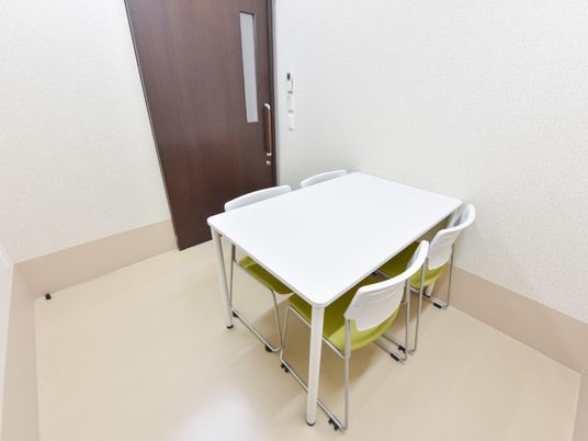 白い壁に囲まれている。壁に寄せて白い四角い4人掛けテーブルがある。椅子の座面は黄緑色、背もたれは四角く白色である。部屋の戸は木目調である。