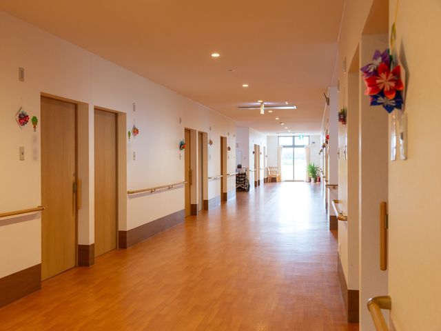 廊下の明るく広々とした空間 共用廊下 明るくゆったりした通路が広がっており、手すりが設置され安全に配慮されています。壁に飾られた工作品が温もりを添え、心地よい環境でのお仕事が想像できます。