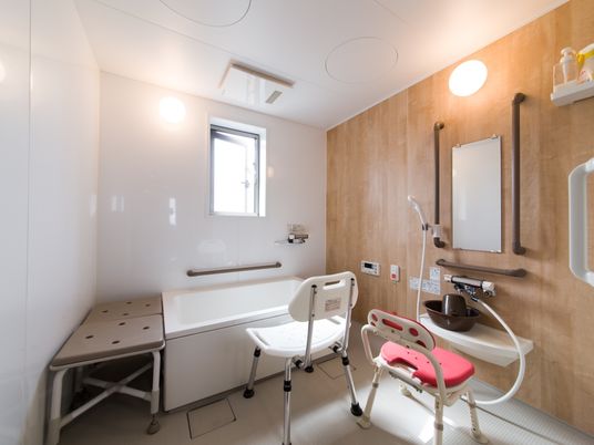 施設の写真 高めの位置に窓があり明るい。浴槽で使用する椅子が２脚用意されている。壁にはナースコールのボタンがある。