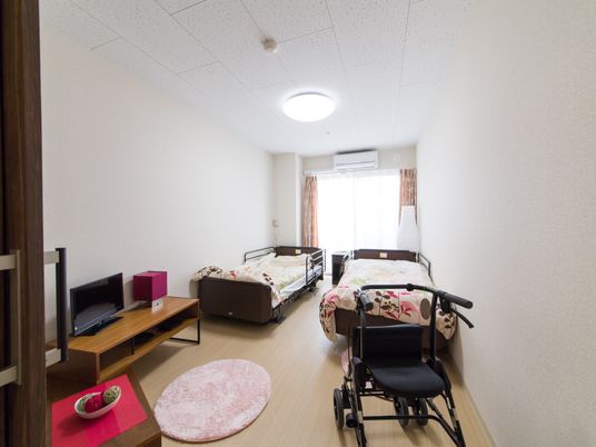 部屋の奥には大きな窓がある。両壁にはベッドが置かれ、手前には車椅子、テレビが置かれている。床には長方形のピンクのマットがある。