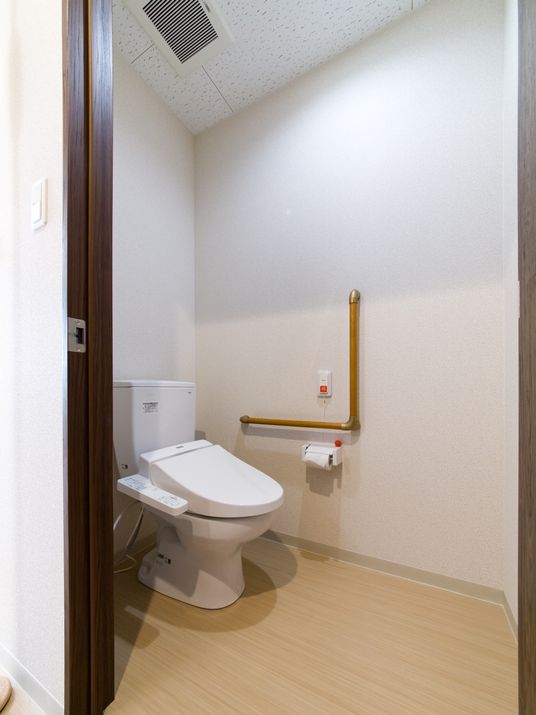 扉が大きく広いトイレ。壁にはＬ字型の手摺が設置されている。緊急用の呼出ボタンもある。温水洗浄機能が付いている。