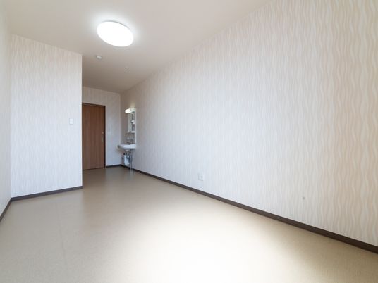 施設の写真 広々とした空間になっている。床はベージュで壁は薄く模様の入った白系になっている。部屋の扉の前に洗面台がある。