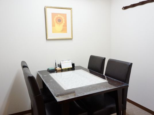 施設の写真 応接室は壁に絵画が飾られ机が1つあり対面で椅子が4つ置かれている。テーブルにはカレンダーやペンなど備品が常備されている。