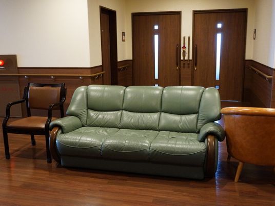 施設の写真 通路には緑色のソファが1つと茶色の椅子が2つ置かれている。壁には手すりと消火栓が設置されており、後ろには通路へと続くドアが見える。