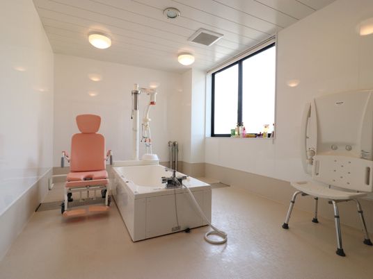 浴室の中央にバスタブがついており、右側の大きな窓からは光が差し込んでいる。介護用のピンク色の車椅子や特殊な機材が並んでいる。