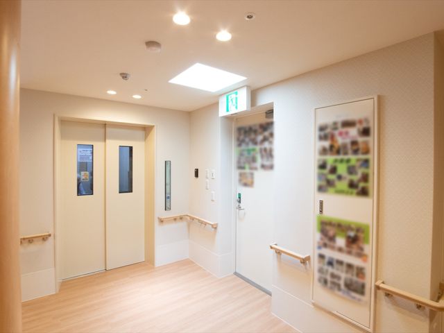 廊下とドアの明るい空間 エレベーターホール ゆとりある廊下は手すりが備えられ、安全に配慮された設計です。壁にはコミュニティの様子が映し出され、温かな雰囲気が漂います。
