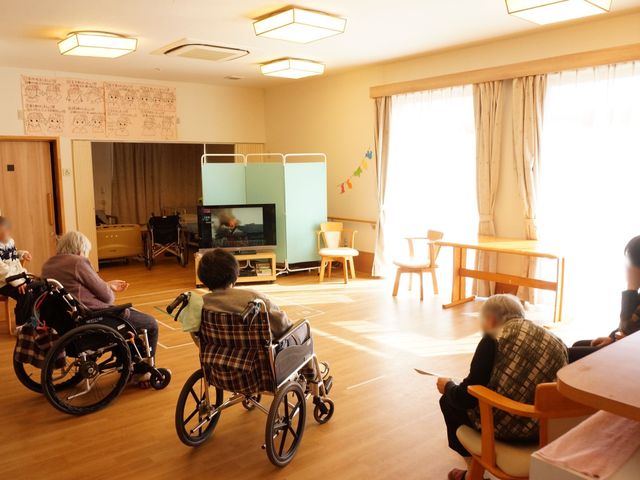 車椅子の利用者室内 サロン 椅子に座ってのんびり過ごす利用者様がいる共有スペースです。日差しが降り注ぐ落ち着いた空間で、ゆったりとした時間をお過ごしいただけます。