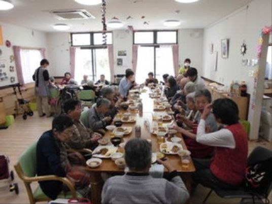 手前から奥にかけて長く繋げられたテーブルで入居者様が食事をしている。左奥にもテーブルがあり、そちらにも数人の入居者様が座っているのが見える。