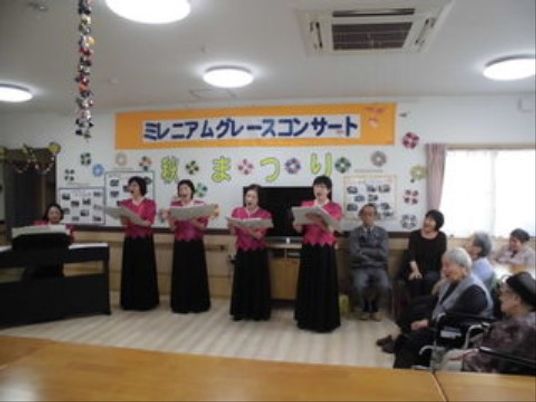 「秋まつり」の幕が飾られた壁の前で、ドレス姿の４人の女性が歌を歌っている。右側に数人の利用者が座って歌を聞いている。