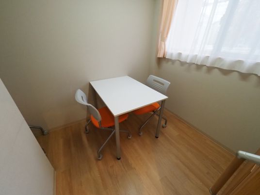 極めてシンプルな応接室の室内。座席は事務用品のようなテーブルと椅子が対面する２人掛けの物が用意されている。