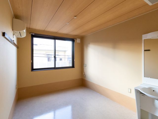 明るい居室の一角 居室 天井まで続く明るい木目調の壁が、居心地の良い個室空間を演出しています。窓からは自然光が差し込み、ゆったりとした時間を過ごせます。