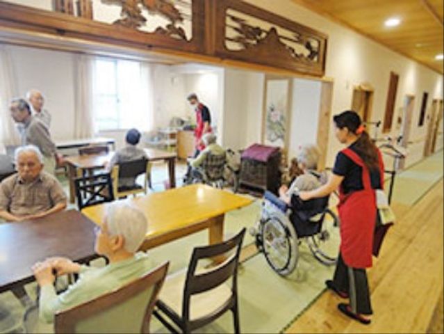 スタッフと入居者の光景 食堂 ゆとりあるダイニングでスタッフが笑顔で対応しています。椅子についてくつろぐ方々の姿も見られます。