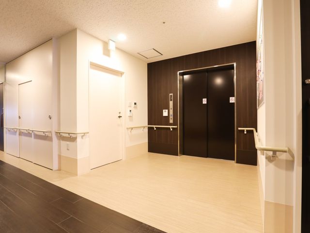 廊下の手すり付き エレベーターホール 落ち着いた色合いでまとめられた廊下は、手すりが設けられており、歩行時の安全を支援しています。壁面の掲示物も、日々の活動に彩りを添えています。