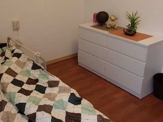 施設の写真 白い壁と茶色のフローリングルームである。３段の白色チェスト、手すりの付いたベッドが置かれており、チェストの上には植物などが飾られている。