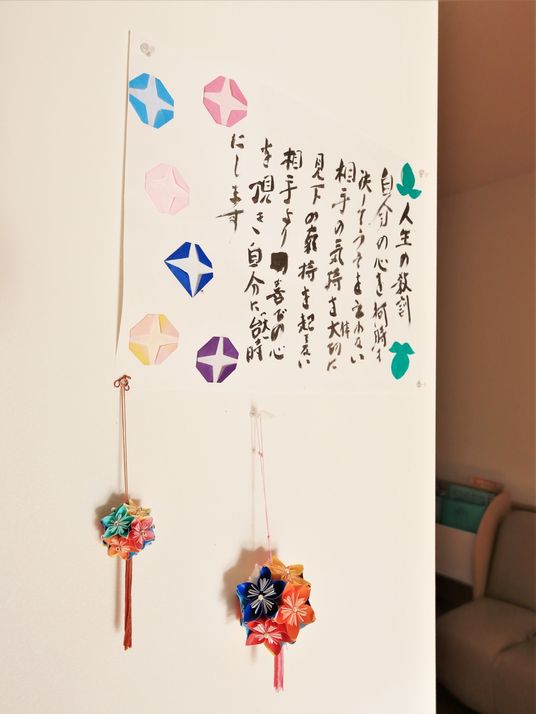 白い壁があり、壁には人生の教訓の文章が書かれた紙が貼られている。周りには折り紙で折られた朝顔や花のくす玉が飾られている。