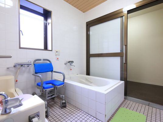 施設の写真 浴室は引き戸で、洗い場と浴槽が分かれている。窓の下には介護用の椅子が置かれており、呼び出しボタンが設置されている。