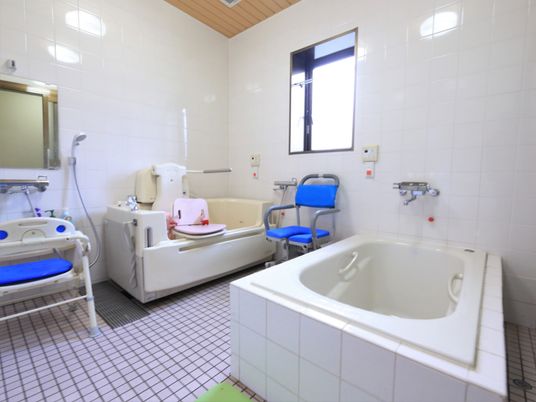 施設の写真 浴室には窓、洗い場の壁には鏡がある。また、介護用の椅子が用意されている。浴槽のほか、介護用入浴機器も設置されている。