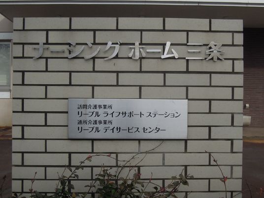 白い壁に施設名が書かれている。下のほうには訪問介護やデイサービスセンターの表記もある。壁の下には花が植えられている。