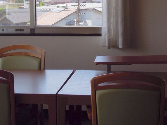 木目調のテーブルが並べられており、椅子が向かい合って置かれている。窓には白いカーテンが掛かっている。
