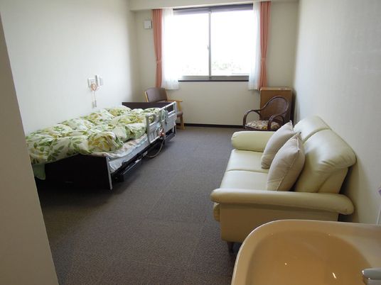 明るい居室は壁が白く、床には茶色のカーペットが敷いてある。クリーム色のソファにはクッションが２個置かれている。