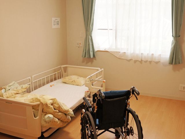 ベッドと車椅子の居室 居室 広々とした居室には日差しの優しい光が差し込み、リラックスできる環境が整っています。車椅子へのアクセスも考慮した配慮が感じられます。