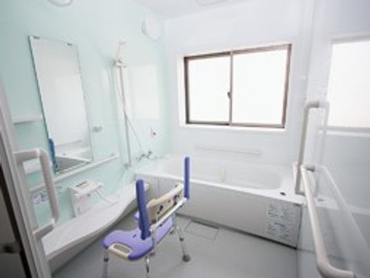 施設の写真 浴槽、シャワー、カラン、鏡、カウンターの付いたユニットバス。壁には窓がある。手すりが設置されており、シャワーチェアが置かれている。