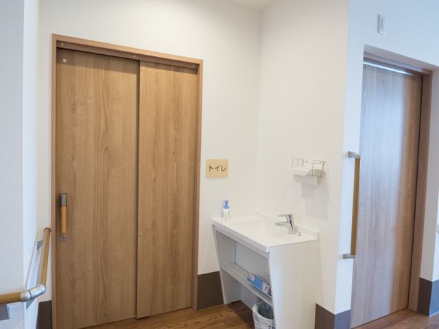 清潔な洗面台エリア トイレ前 手すり付きで安全性を考えた洗面所は、ご利用者の便宜を図っています。木目調の扉が温もりを感じさせる空間です。