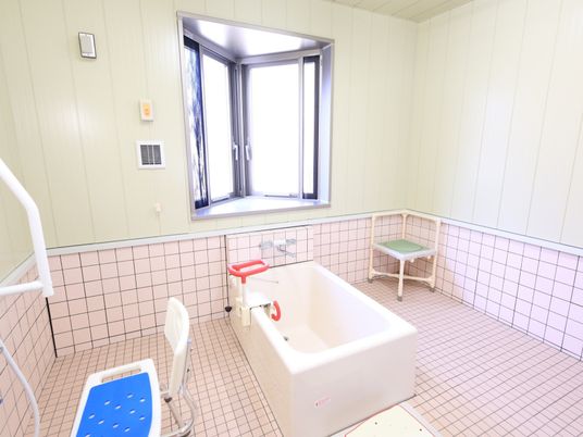 出窓が設置された広々とした浴室である。ピンク色のタイルが床と壁に貼られ、洗い場に青いシャワー椅子が置いてある。