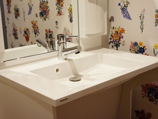 施設の写真 カラフルな大きい花柄の壁紙が印象的な洗面所である。車椅子をご利用の方も手を伸ばしやすい洗面台が使われている。