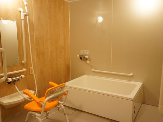 施設の写真 広い洗い場スペースがある浴室。壁には鏡や手すりが設置してあり、ひじ掛けと背もたれがついたシャワーチェアが用意してある。