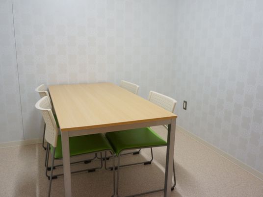 施設の写真 白の壁紙にベージュの床である。相談室のような部屋である。椅子と机が置かれている。落ち着いた部屋で話しができる。