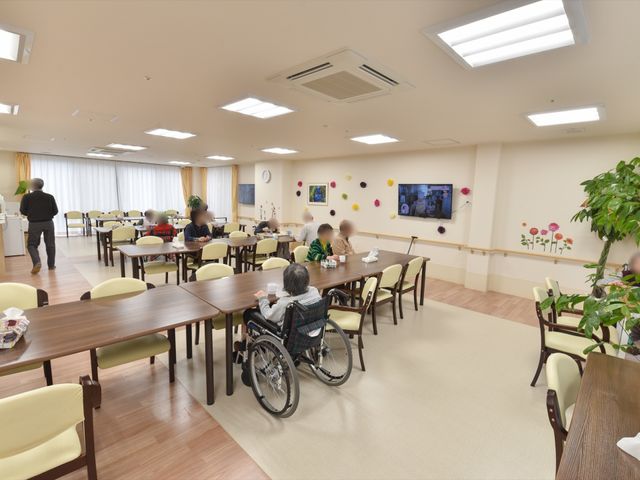 利用者が食事する食堂 食堂 ゆったりとした空間で利用者が食事を楽しんでおり、壁に飾られた花が明るい雰囲気を演出しています。テレビもあり、食事時だけでなく憩いの時間も充実していそうです。