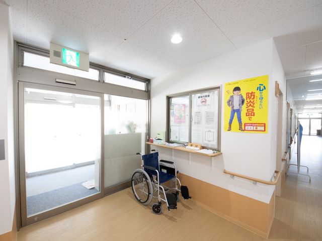 車椅子の置かれた廊下 受付 ゆったりとした廊下には掲示物があり、利用者の安全と情報提供に配慮された空間が広がってます。車椅子も配置され、バリアフリーな設計が工夫されています。