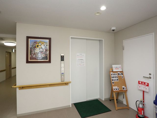 廊下に掲示物とエレベーター エレベーターホール 手すり付きの廊下には明るい絵画が飾られ、利便性を考えたエレベーターが配されています。入居者の安全と快適な移動を支える設計です。