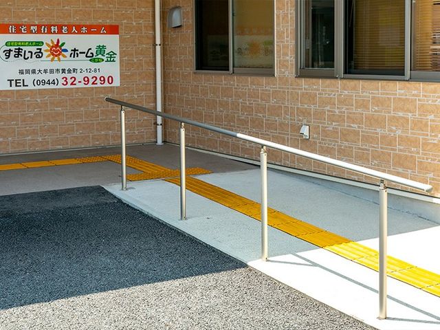手すり付きの出入口 外観   手すりと段差を抑えた通路があり、車椅子でも移動しやすい出入口です。壁面には案内看板が設置されています。