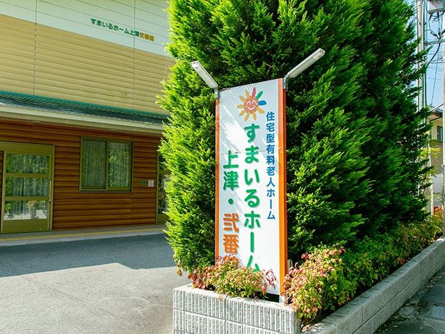 施設看板と建物外観 外観   緑の植栽のそばに看板が立ち、落ち着いた雰囲気の建物外観です。日差しが入り、出入口まわりも明るく見えます。