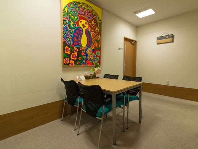 カラフルな絵とテーブルの休憩室 相談室 落ち着いた空間の休憩室では、鮮やかな壁画を眺めながら、ゆったりとくつろぐことができます。明るい木目調のドアが、清潔感ある印象を与えています。