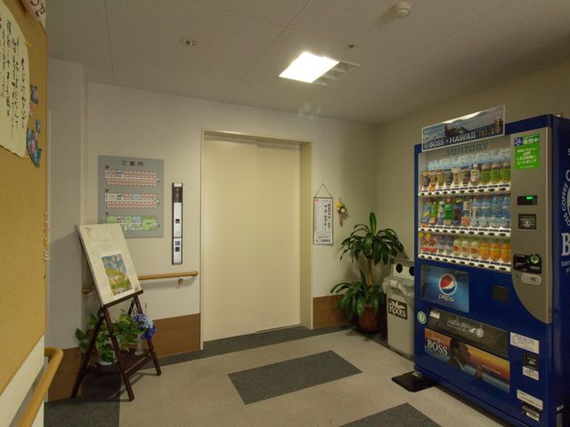 廊下と自動販売機 エレベーターホール ゆったりした廊下に、利用者の憩いのスペースとして設置された自動販売機が見られます。壁には掲示物も飾られ、明るく和やかな雰囲気を感じられます。