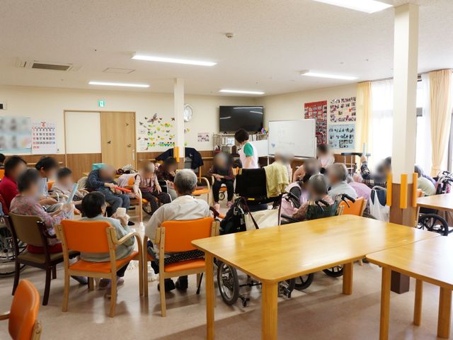 多目的利用の広間 多目的室 共有する広い空間で、入居者が様々な催しに参加しています。スタッフが笑顔で対応し、温かな雰囲気が漂っています。