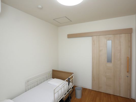 木目調のスライドドアがあり、壁際にベッドが置かれている。広めのスペースがある。天井にはシーリングライトが設置されている。