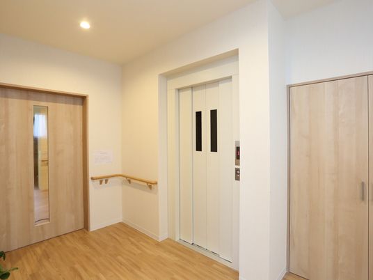 真っ白な扉のエレベーターがある。横の壁には手すりが設置されていて、収納庫や別の部屋の扉が見えている。
