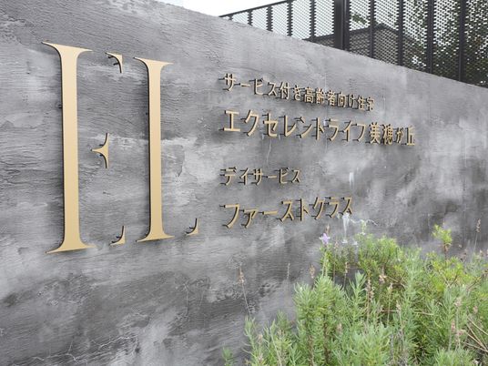 灰色のコンクリートの壁に、金色の鉄製の文字型のプレートで施設名が表示されている。壁の前は植え込みになっており、草木が植えられている。