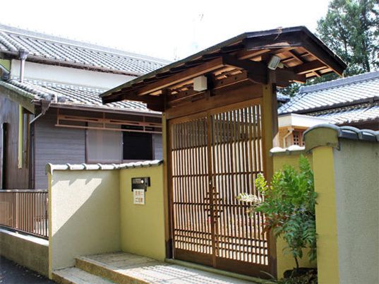 施設の写真 伝統的な日本家屋のような数寄屋門があり、その先に瓦屋根の建物がある。門の前には２段の段差があり、門の横には植物がある。