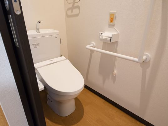 施設の写真 白を基調とした清潔なトイレである。洗浄機能付きの便座が設置されている。壁には手すりとナースコールが取り付けてある。