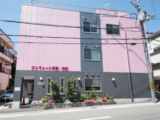 施設の写真 ピンク色の目立つ３階建ての建物となっている。建物の２階部分の壁にに大きく赤い文字で施設名が入れてある。施設の横に花壇が設けてある。