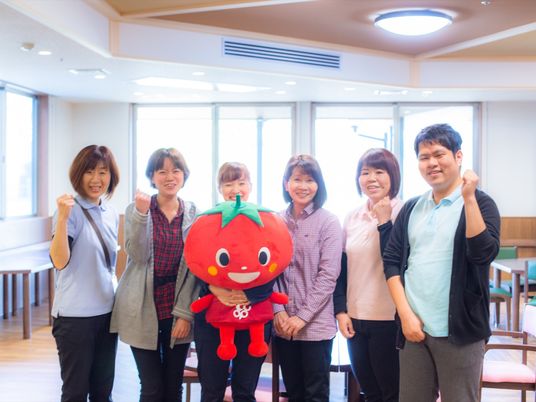 施設の写真 リビングを背景に、六人の男女のスタッフが笑顔で写っている。マスコットキャラクターのぬいぐるみを持っている。
